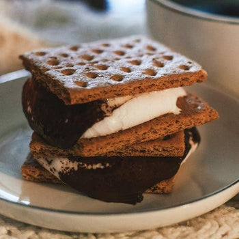 Homemade keto graham crackers stacked on a wooden board with cinnamon sticks, almond flour, butter, and a cup of coffee beside them.